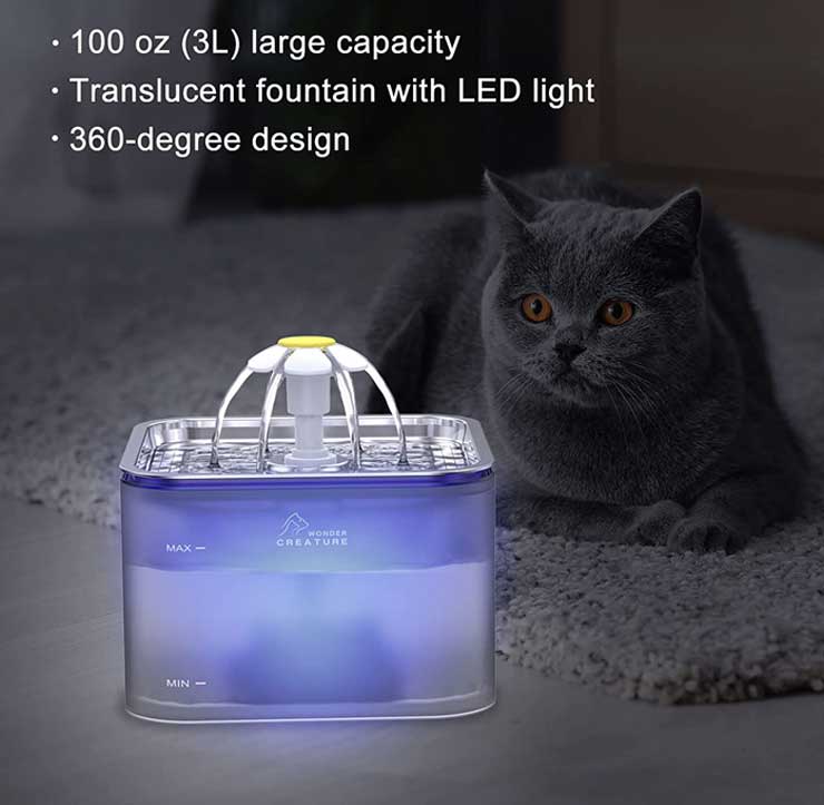 A dark cat is lying next to a cat water fountain in dark blue color with LED lights